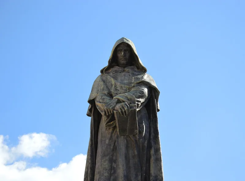 En mørk bronsestatue av en munk med hette og en bok i hendene, sett nedenfra mot blå himmel.