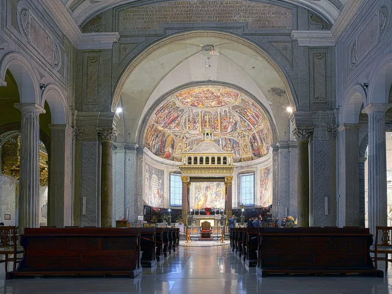 Midtskipet i en basilika med antikke søyler på begge sider, trebenker og en freskomalt apsis med baldakin over alteret.