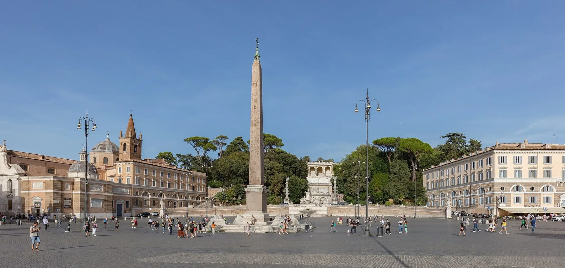 Et stort, åpent ovalt torg med en egyptisk obelisk i midten, omkranset av kirker, lyse bygninger og trær under blå himmel.