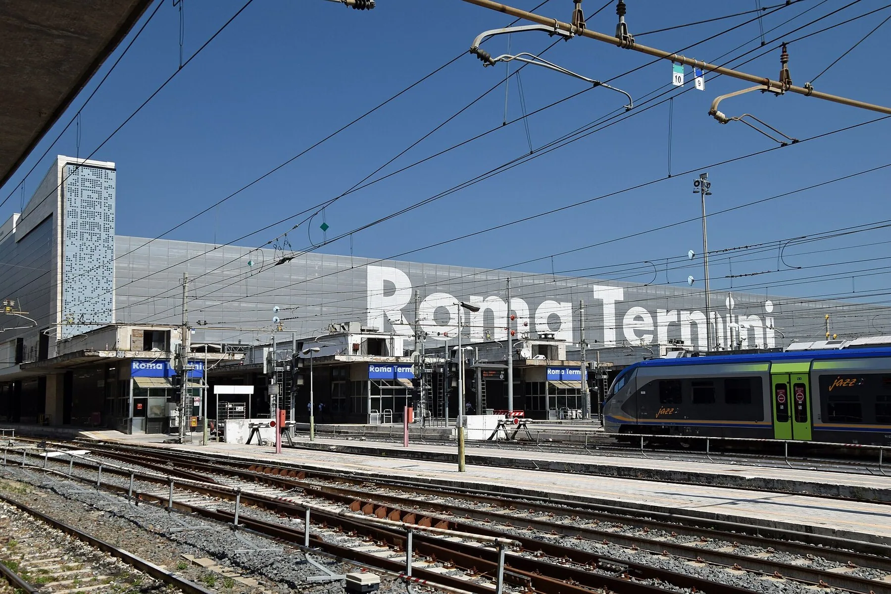 Jernbanespor og plattformer foran en lang stasjonsbygning med stor hvit skrift på fasaden, og et regionaltog til høyre under kjøreledningene.