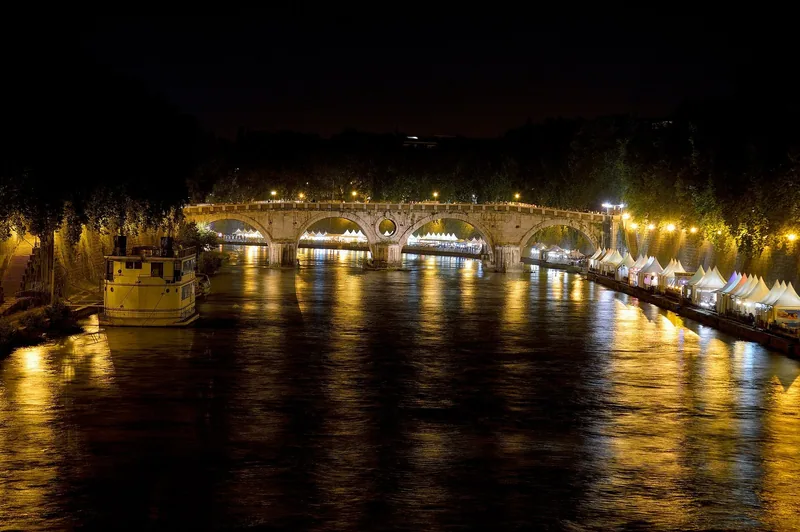 En steinbro med fire buer over en elv om kvelden, med gatelys langs kaiene og hvite telt nede ved vannkanten.