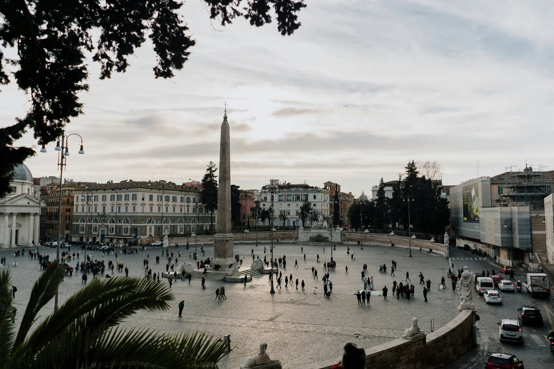 Piazza del Popolo i ettermiddagslys sett ovenfra: obelisken står midt på den store brosteinsplassen, folk går spredt over den, og lyse fasader og trerekker rammer den inn.