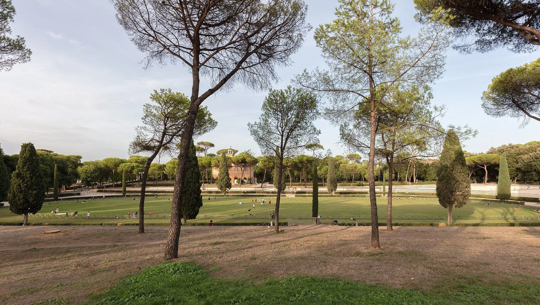 Den store gressflaten ved Piazza di Siena i Villa Borghese, med brede paraplypinjer og smale sypresser i rekke rundt kanten og noen få mennesker som sitter i skyggen under trærne.