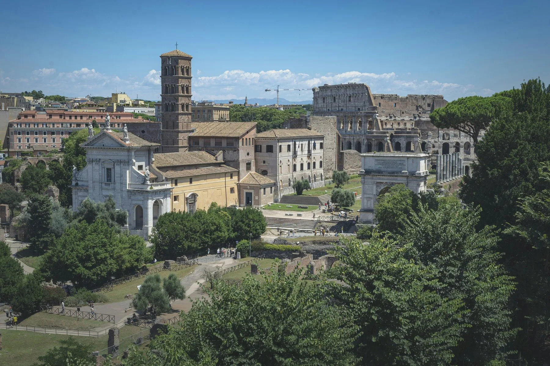 Colosseum sett ovenfra med ruinene og pinjetrærne rundt, og bytak som strekker seg bakover i sommerdisen.