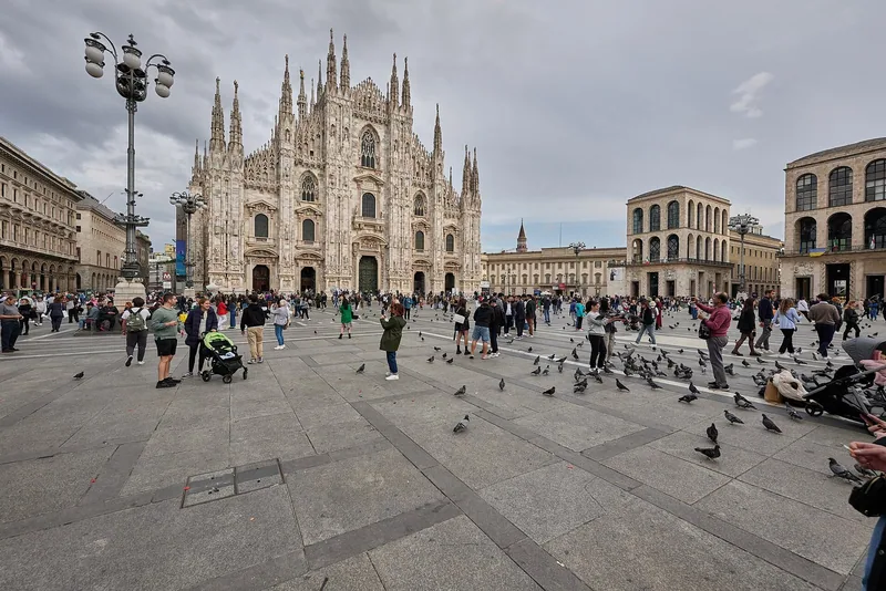 Den hvite marmorfasaden på domkirken i Milano med de mange spirene, sett over Piazza del Duomo med folk og duer.