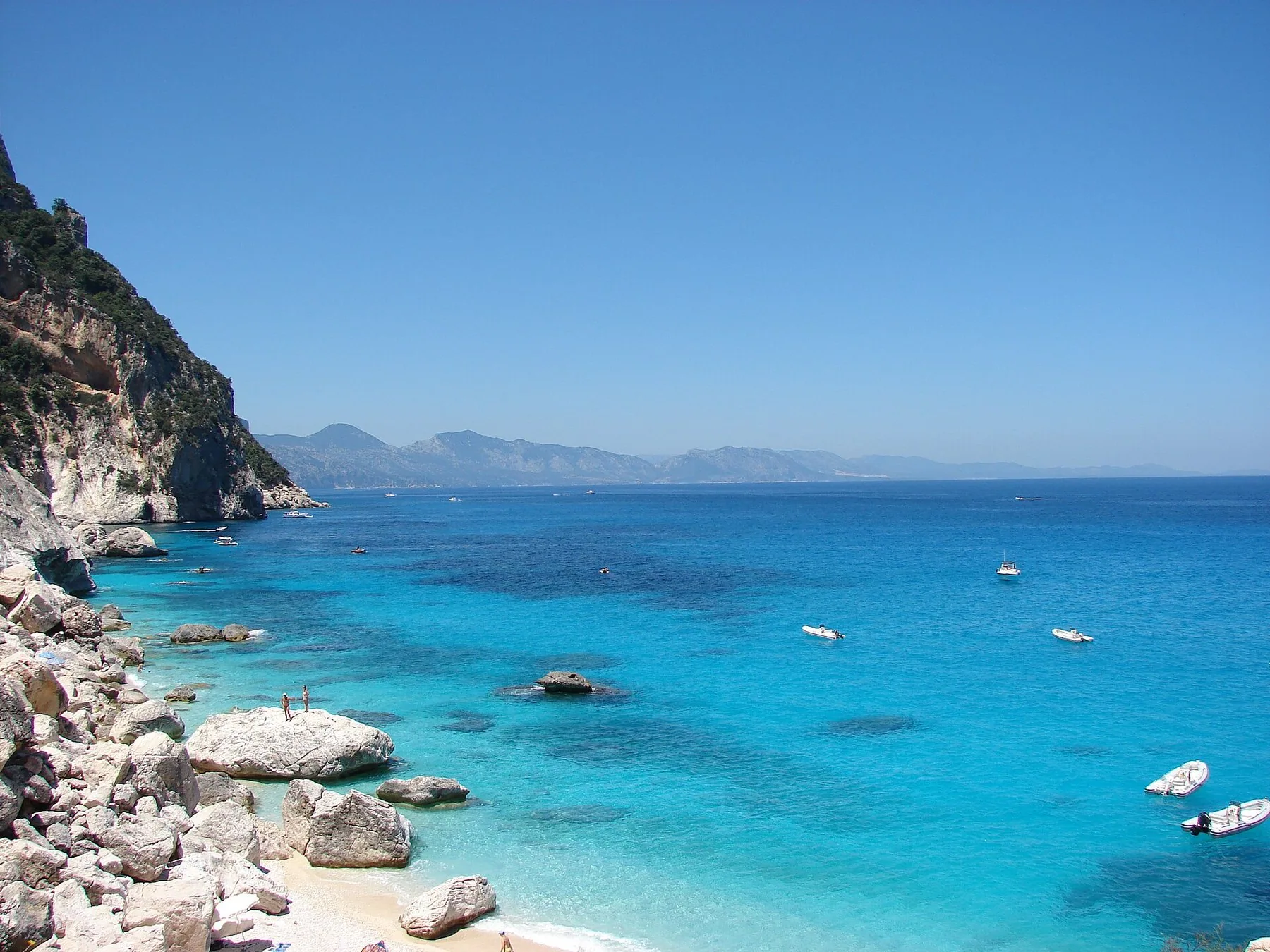 Cala Goloritzé med hvite steinblokker i strandkanten, vann som går fra turkist til dypblått, og noen småbåter som ligger for anker utenfor bukta.