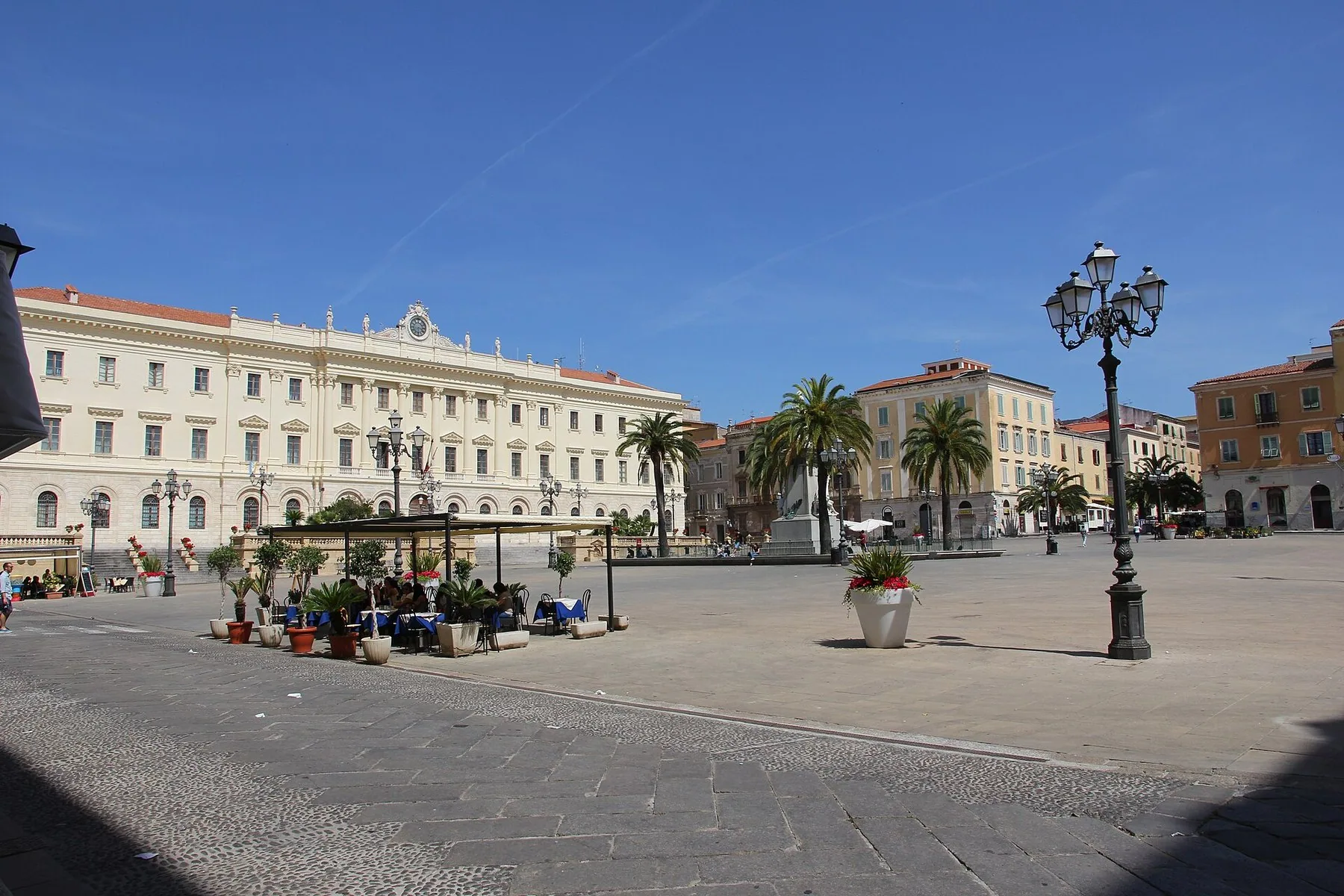 Piazza d'Italia i Sassari, en stor åpen plass med palmer, gatelykter og den lyse fasaden på Palazzo della Provincia under blå himmel.