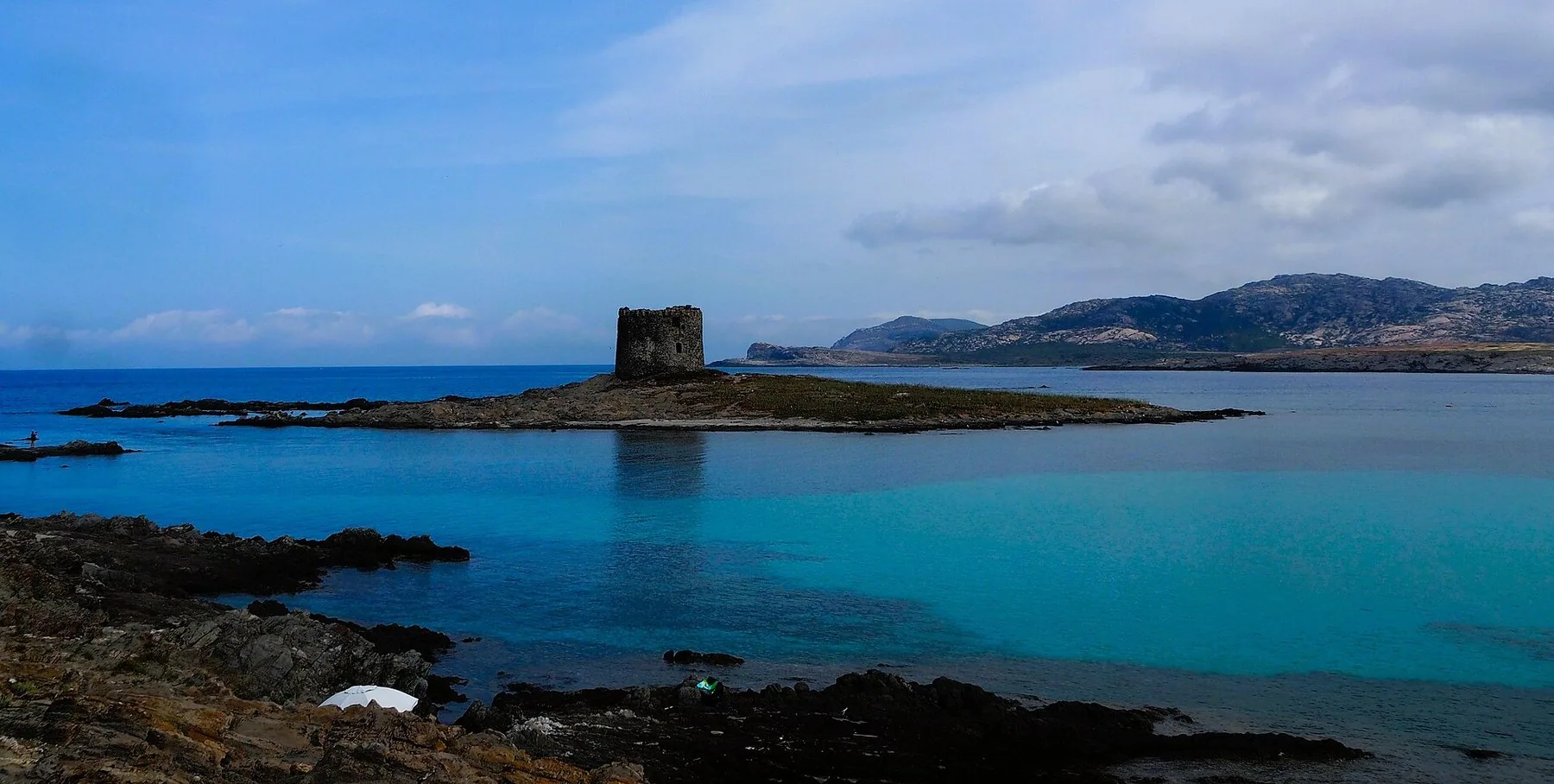 Turkist, grunt vann ved Stintino med det runde vakttårnet Torre della Pelosa på en lav holme og lave åser i bakgrunnen.