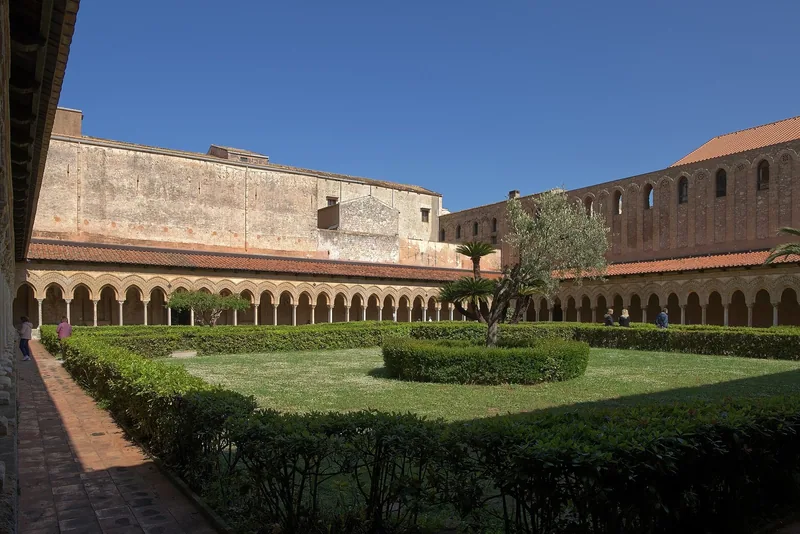 Klostergården i Monreale med lave buerekker rundt en klippet hage, et oliventre i midten og høye murer under blå himmel.