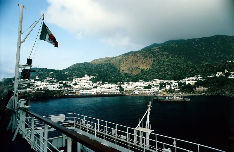 Hvite hus langs kysten av Panarea sett fra dekket på en ferge som legger til, med grønne åser bak og et italiensk flagg i vinden.