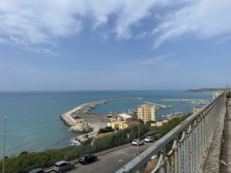 Utsikt fra et rekkverk ned mot havna i Sciacca, med moloer, fiskebåter og en vei langs kysten.
