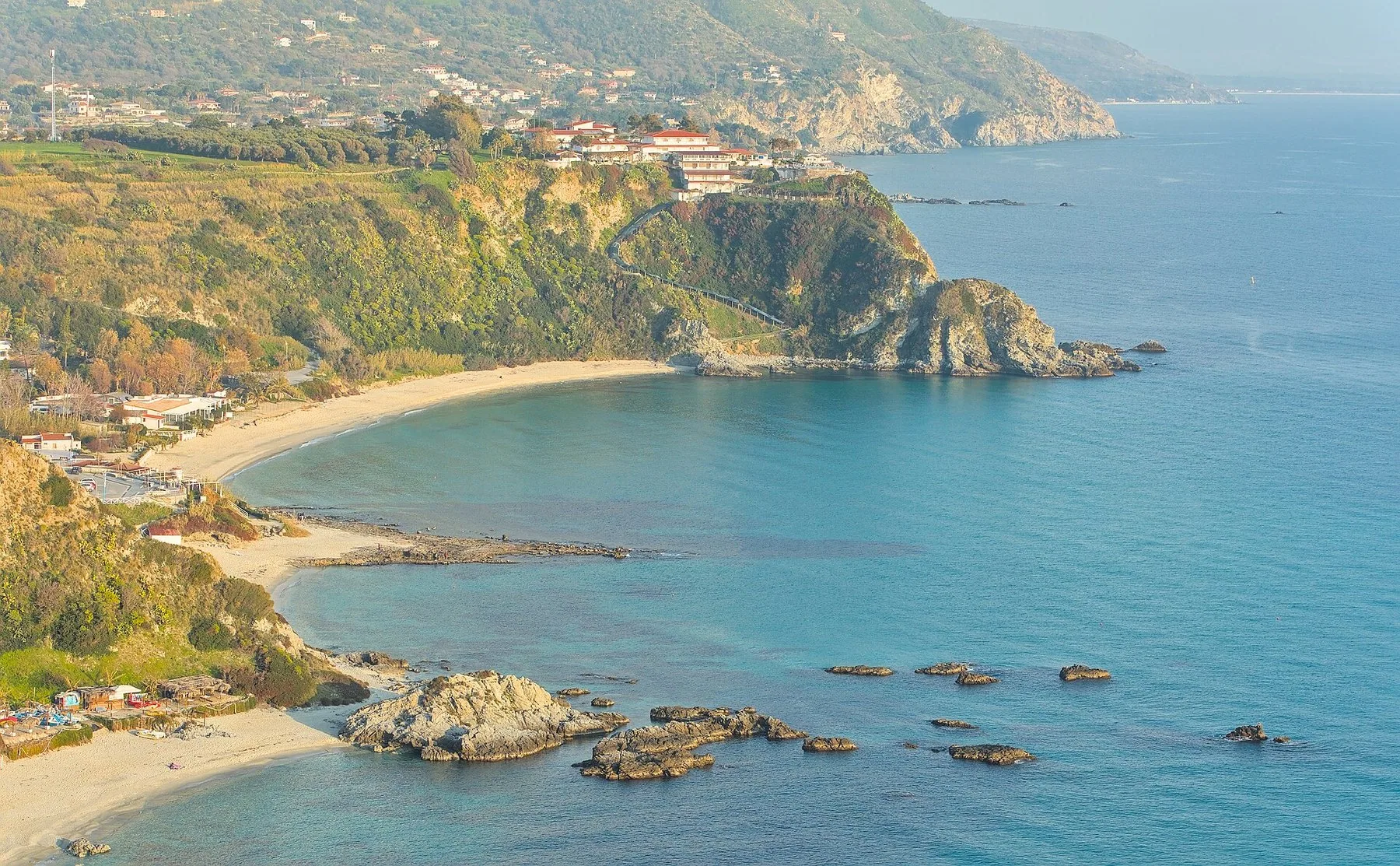 En bue av lys sandstrand ligger mellom to grønnkledde odder på kysten av Calabria, med lave skjær i grunt turkist vann og hvite hus høyt oppe på pynten.