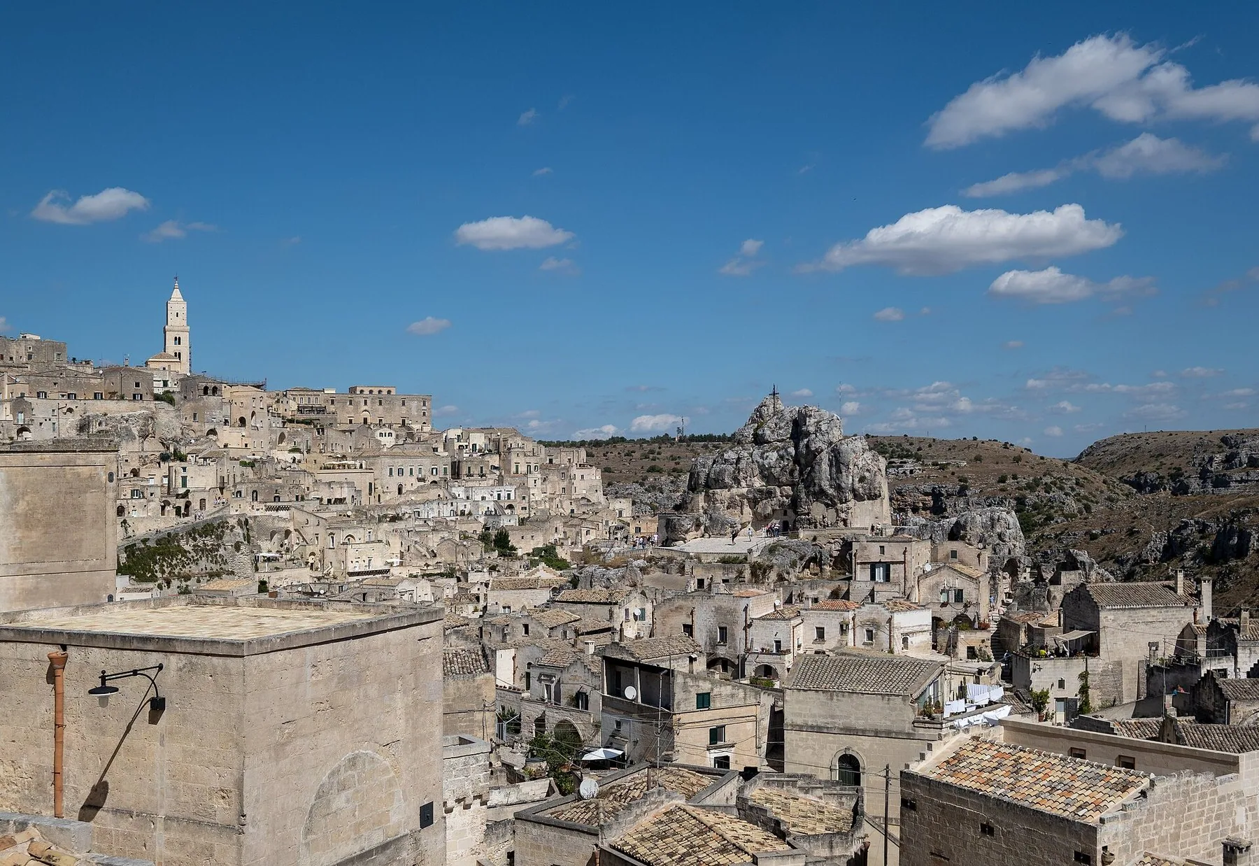 Husene i Matera ligger tett i tett i lys kalkstein oppover skråningen mot et hvitt klokketårn, med en naken klippeformasjon og ravinen til høyre.