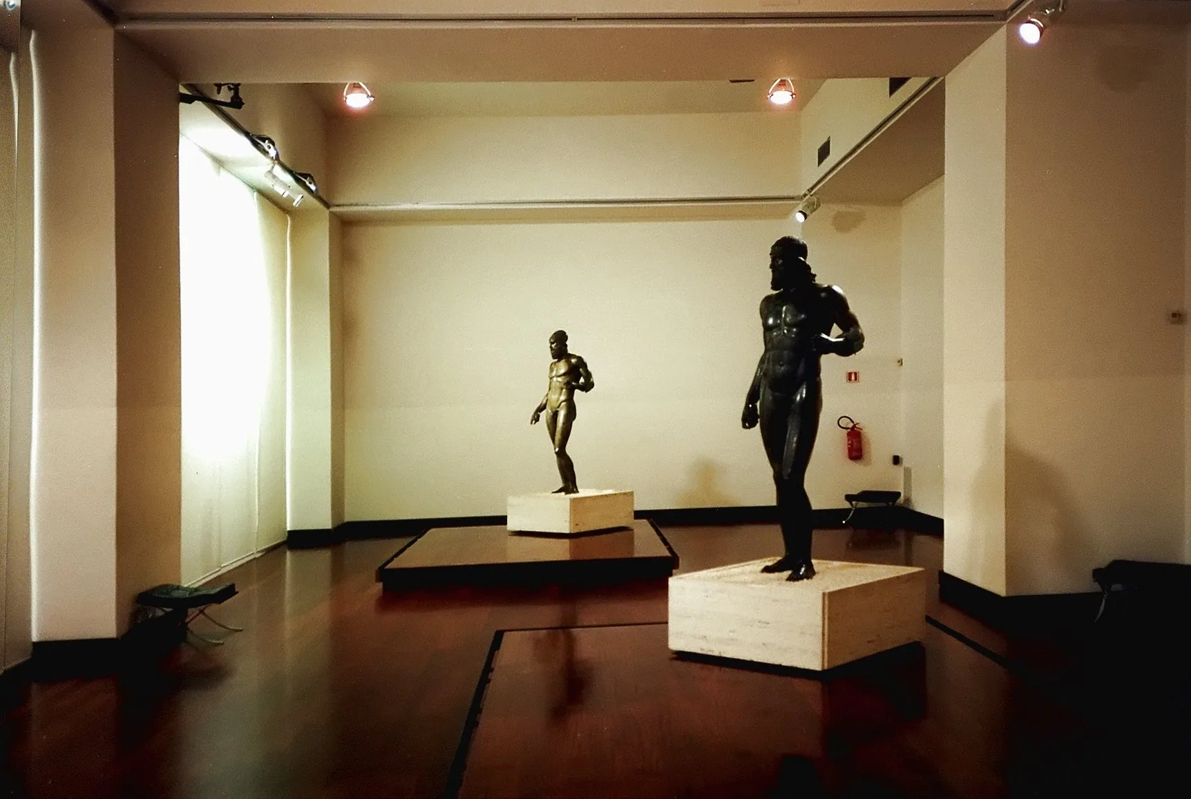 To bronsestatuer av nakne, skjeggete menn står på hver sin lave sokkel i et opplyst museumsrom med mørkt tregulv og lyse vegger.