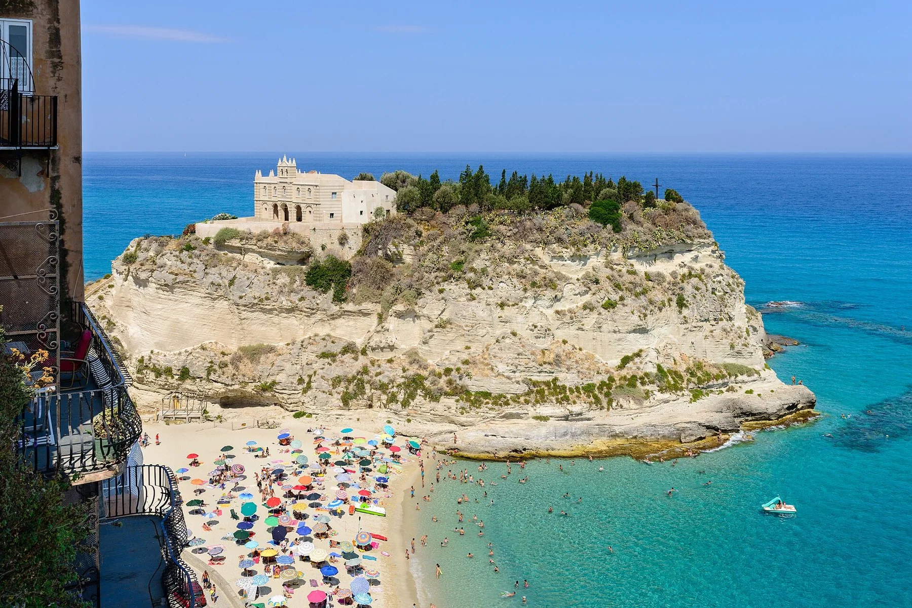 Et fritt liggende klippeskjær utenfor Tropea med et gammelt kloster og noen sypresser på toppen, og en sandstrand full av parasoller og badende i turkist vann foran.