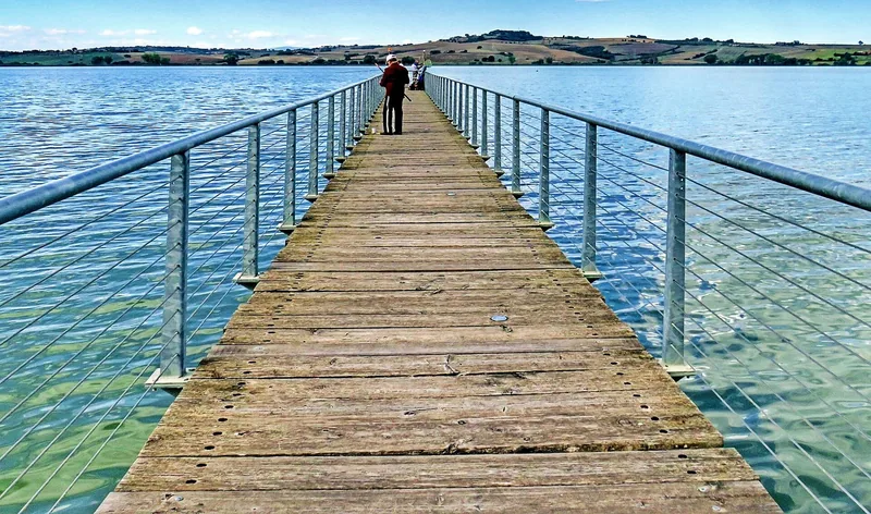 En lang trebrygge med rekkverk går ut i det blå vannet på Lago di Chiusi, med lave, grønne åser på andre siden.