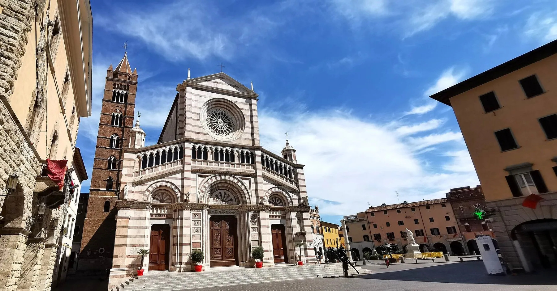 En katedral med stripete marmorfasade, rosevindu og et teglklokketårn ved et solfylt torg under blå himmel med hvite skyer.