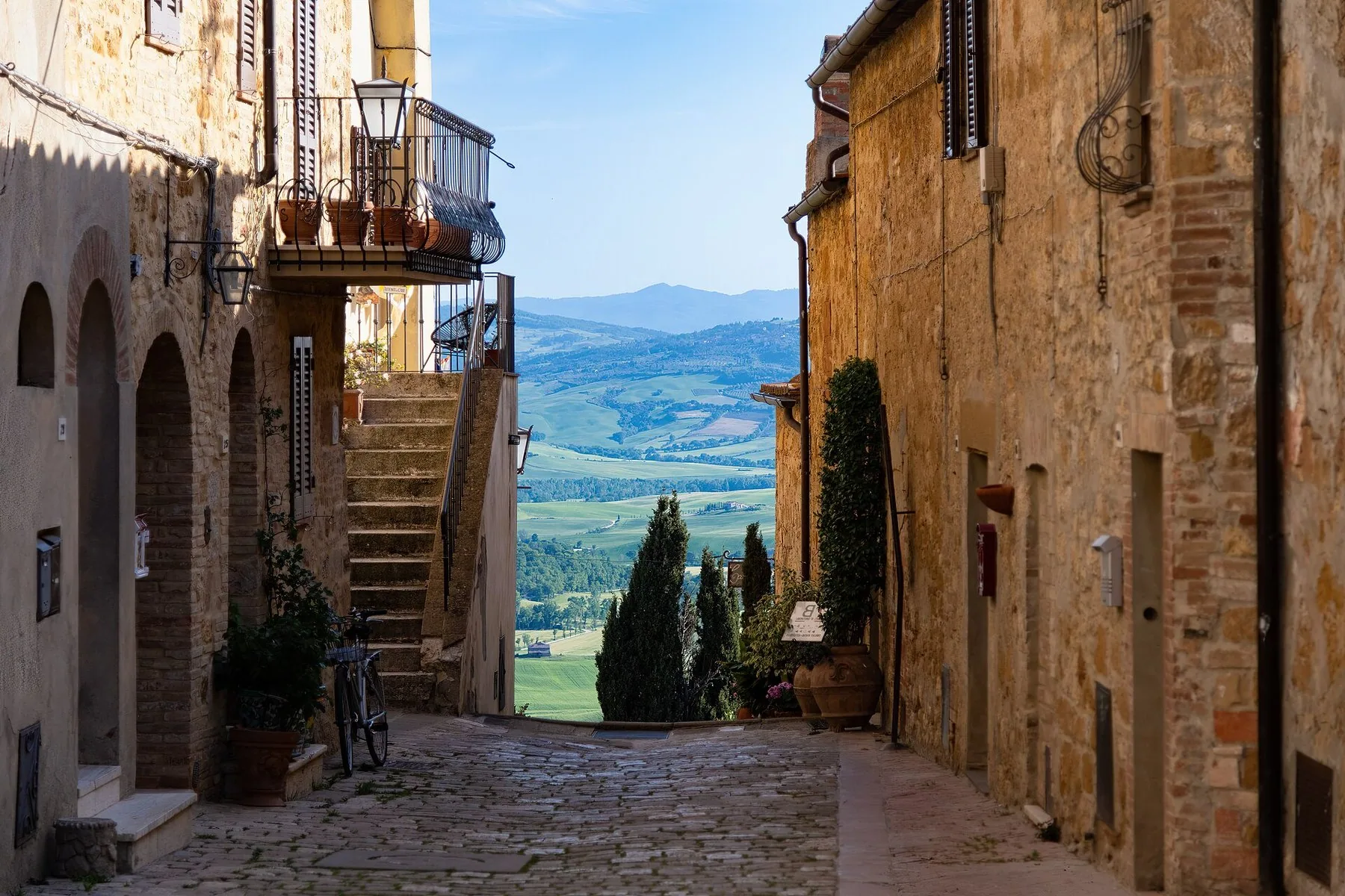 En smal, brolagt gate i Pienza mellom hus i lys stein åpner seg mot dallandskapet nedenfor, med en smijernsbalkong og potteplanter til venstre, en utvendig trapp og en enslig sypress i åpningen.