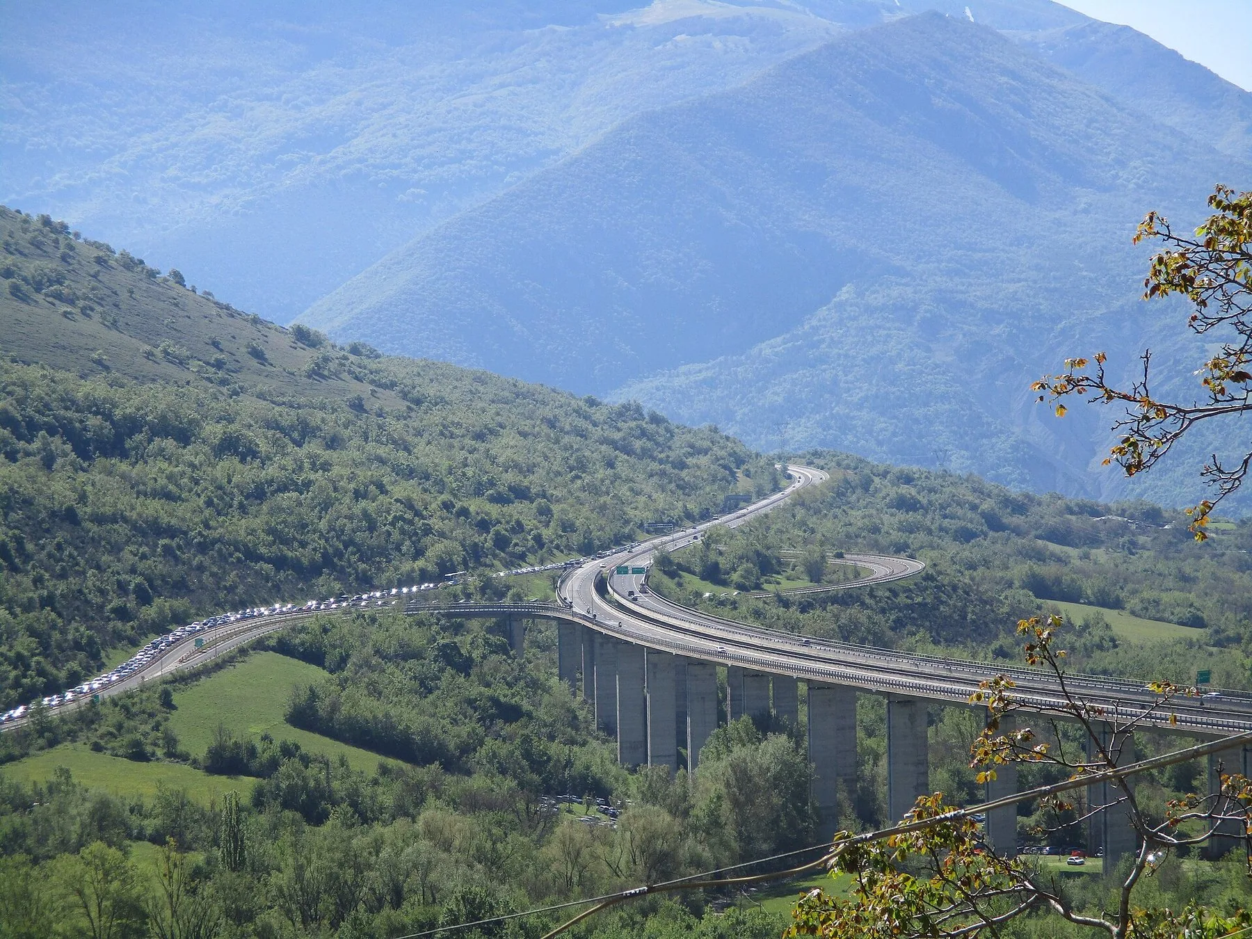 En motorvei på høye betongpilarer svinger gjennom en grønn fjelldal, med en lang kø av biler på den ene kjøreretningen og blånende fjell bak.