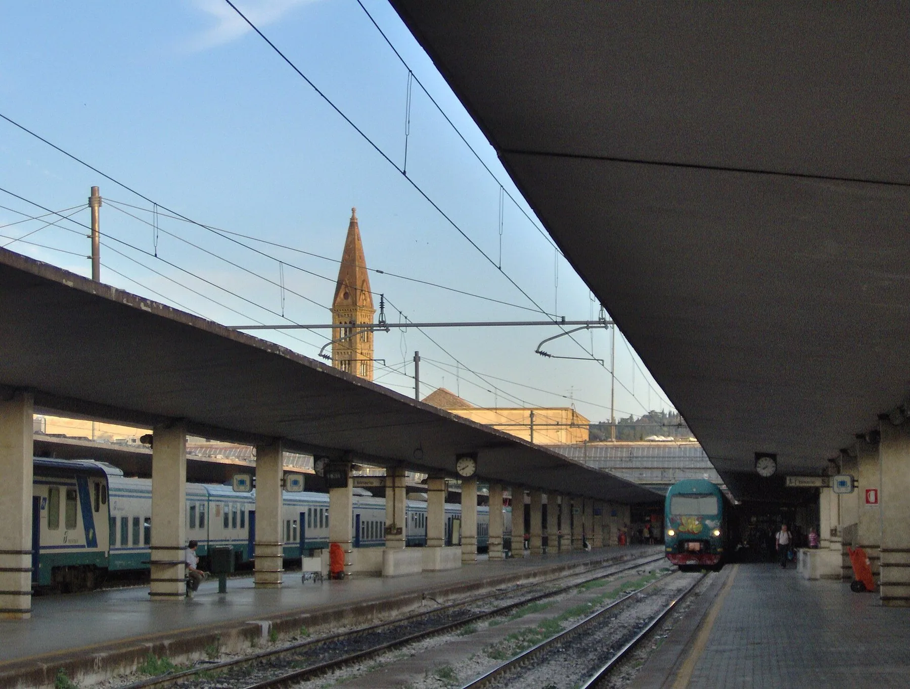 Plattformer med lange betongtak og tog på sporene på Santa Maria Novella stasjon i Firenze, med klokketårnet til kirken Santa Maria Novella i bakgrunnen.