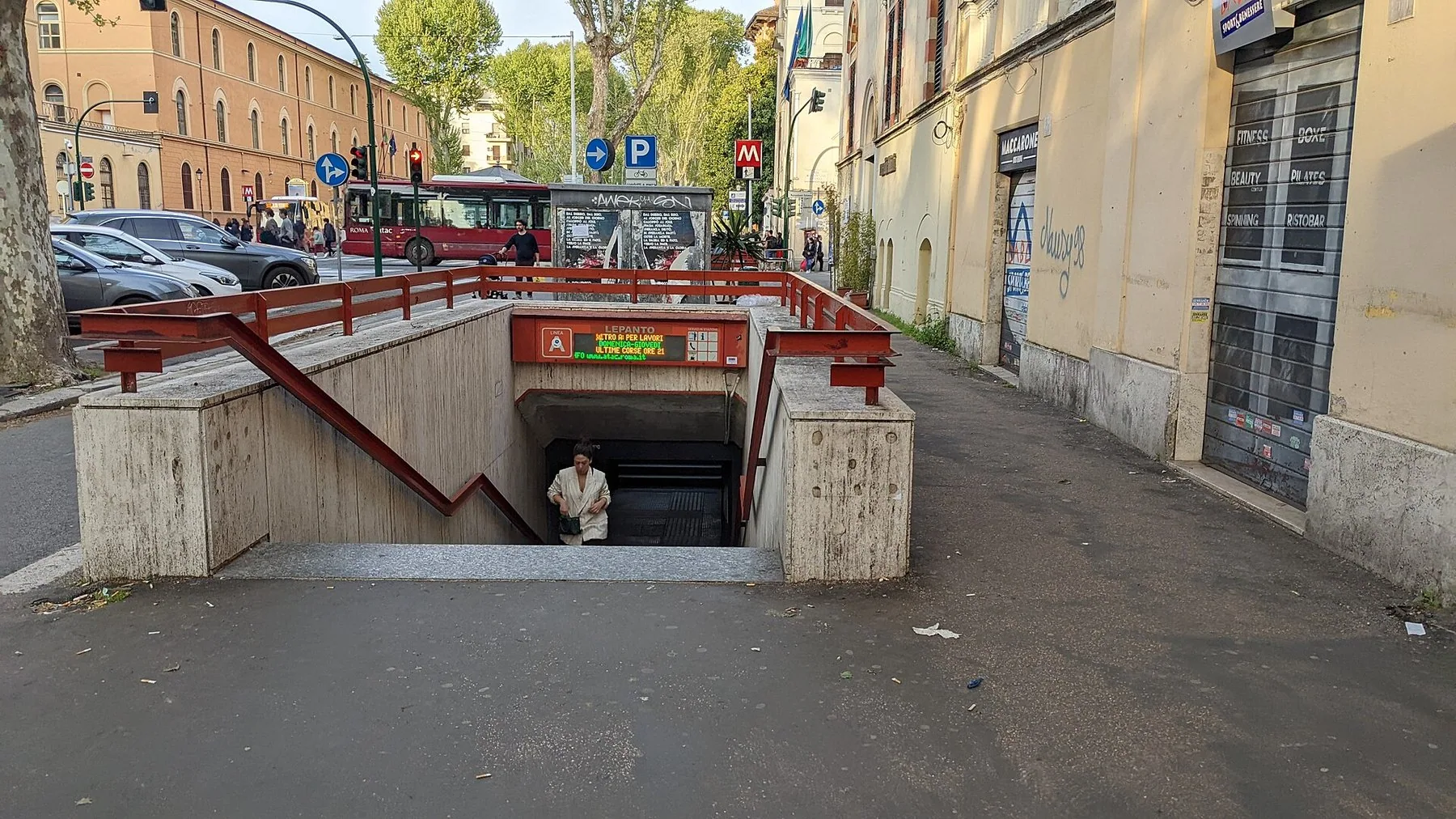 En nedgang til en metrostasjon midt i fortauet, med røde rekkverk, en lystavle over trappen og en rød bybuss som passerer i gaten bak.