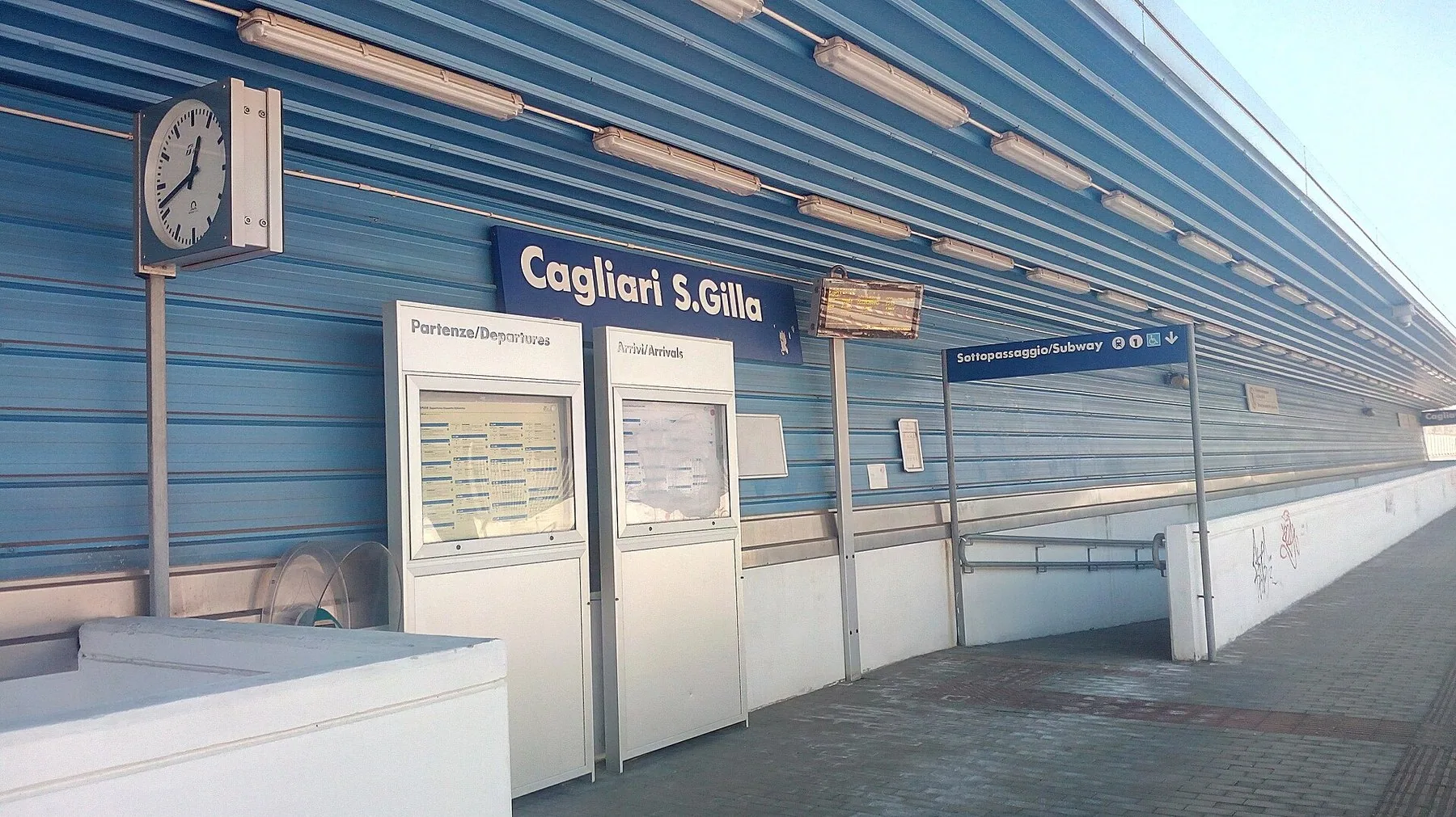 En tom perrong ved en liten stasjon, med et leskur, en klokke, stasjonsskilt og oppslagsskap for avgangs- og ankomsttider langs veggen.