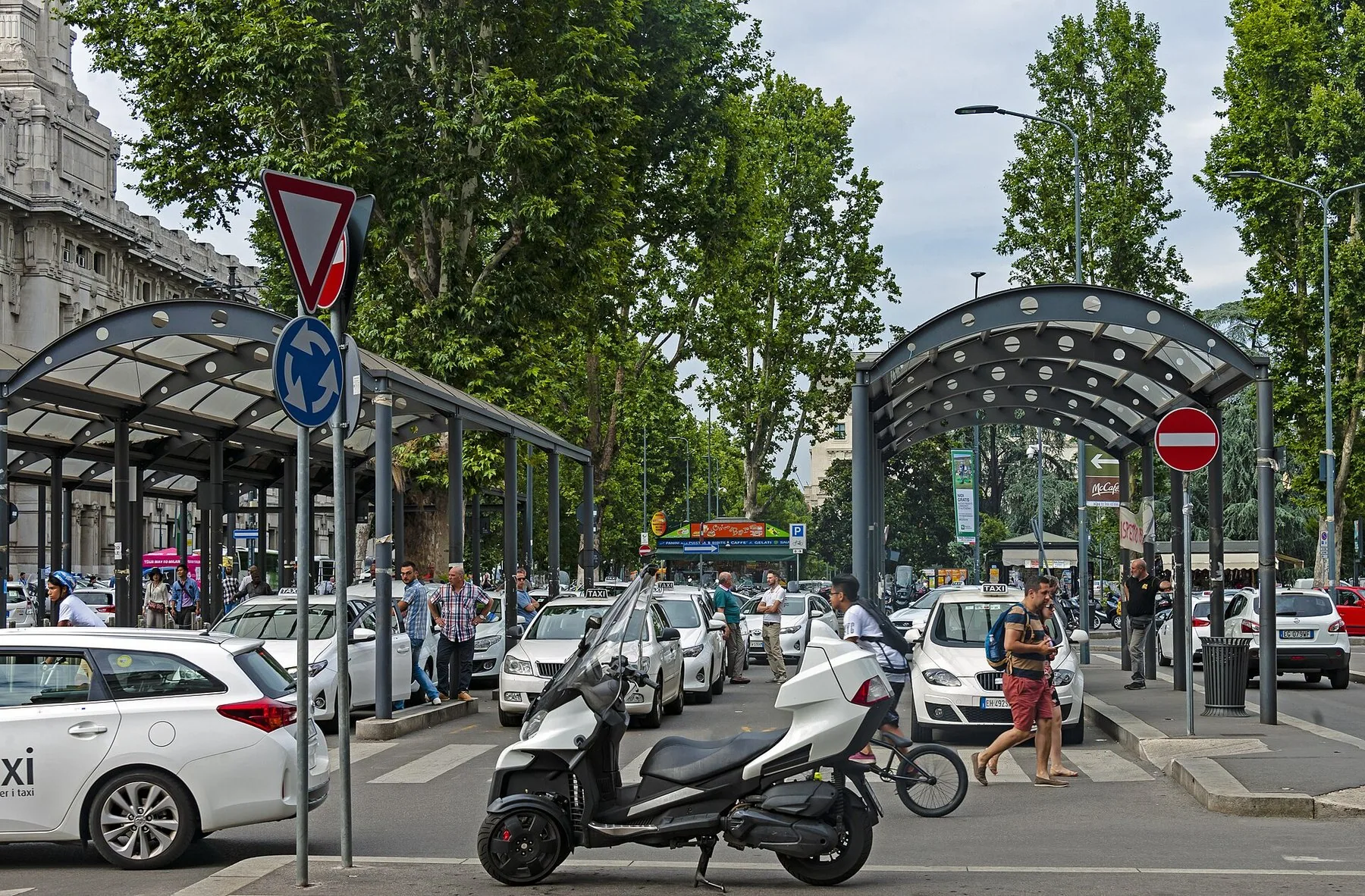En taxiholdeplass med hvite drosjer i kø under buede leskur, med en scooter og fotgjengere i forgrunnen.