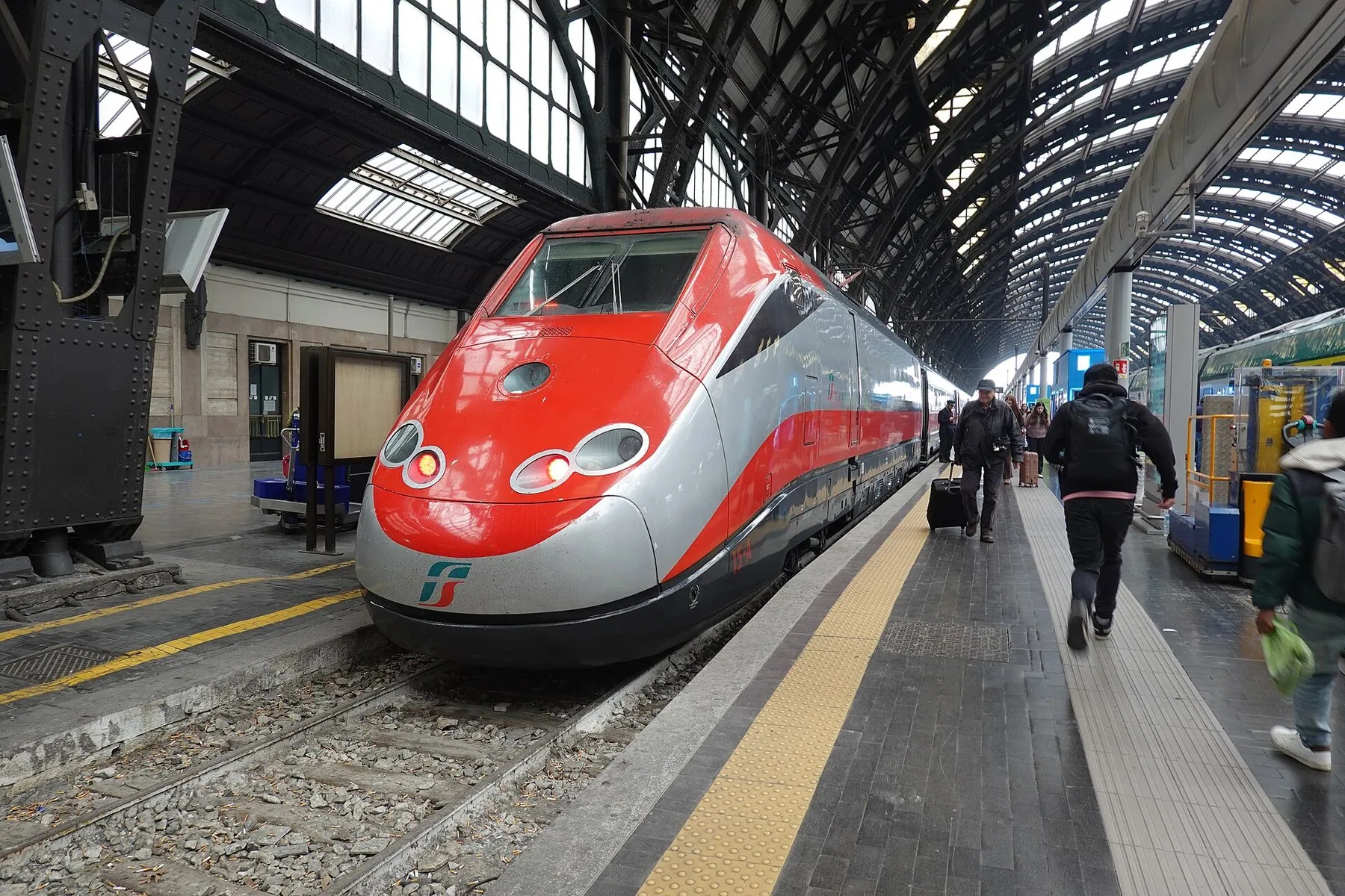 Et rødt og sølvgrått høyhastighetstog står ved plattformen under det buede stål- og glasstaket på Milano Centrale, med reisende som går langs toget.