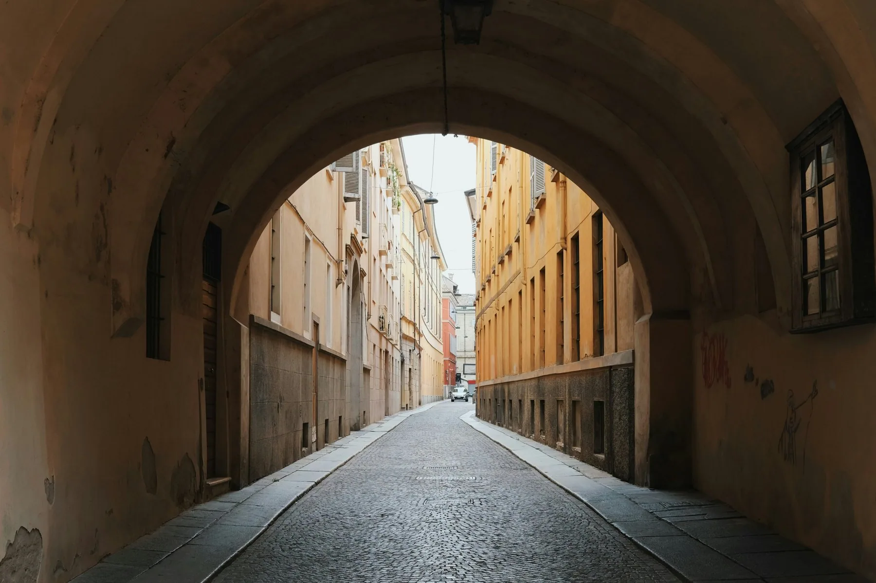 Utsikt gjennom en mørk steinhvelving mot en tom gate med våt, blank brostein og høye husfasader på begge sider.