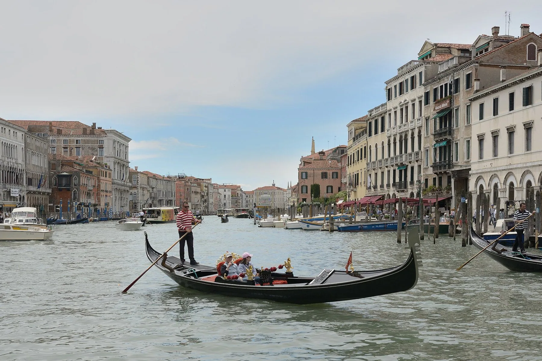 En gondolier står bakerst i den svarte gondolen og støter åren gjennom vannet midt på Canal Grande, med palassfasader og fortøyningsstolper på begge sider.