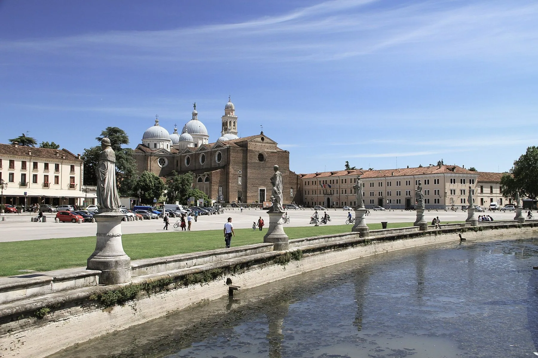 Den ovale kanalen midt på Prato della Valle er kantet av hvite marmorstatuer på sokler, og bak plassen reiser kuppelrekken på Santa Giustina-basilikaen seg mot blå himmel.