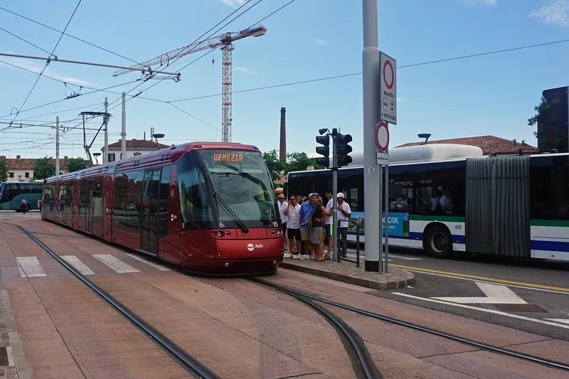 En rød trikk kjører inn på en holdeplass med skinner i bakken, mens en leddbuss står ved siden av og folk venter under kontaktledningene.