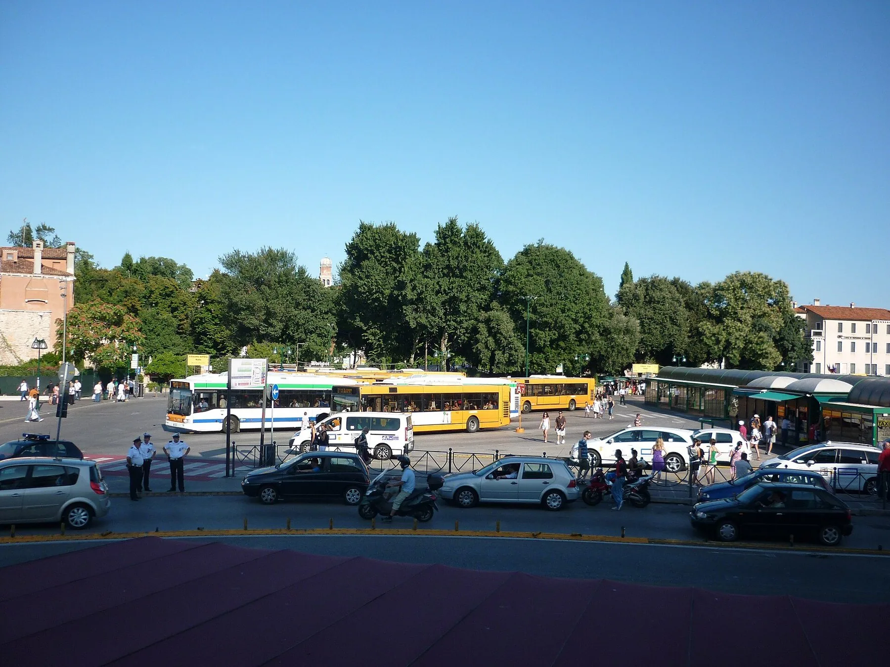 Stor åpen plass med busser, taxier og parkerte biler foran en trerekke, med lave bygninger i bakgrunnen under blå himmel.