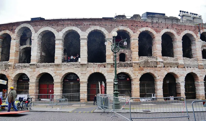 Den romerske arenaen i Verona sett på nært hold, med to rekker rundbuer i rosa og hvit kalkstein og røde inngangsporter nederst mot brosteinsplassen.