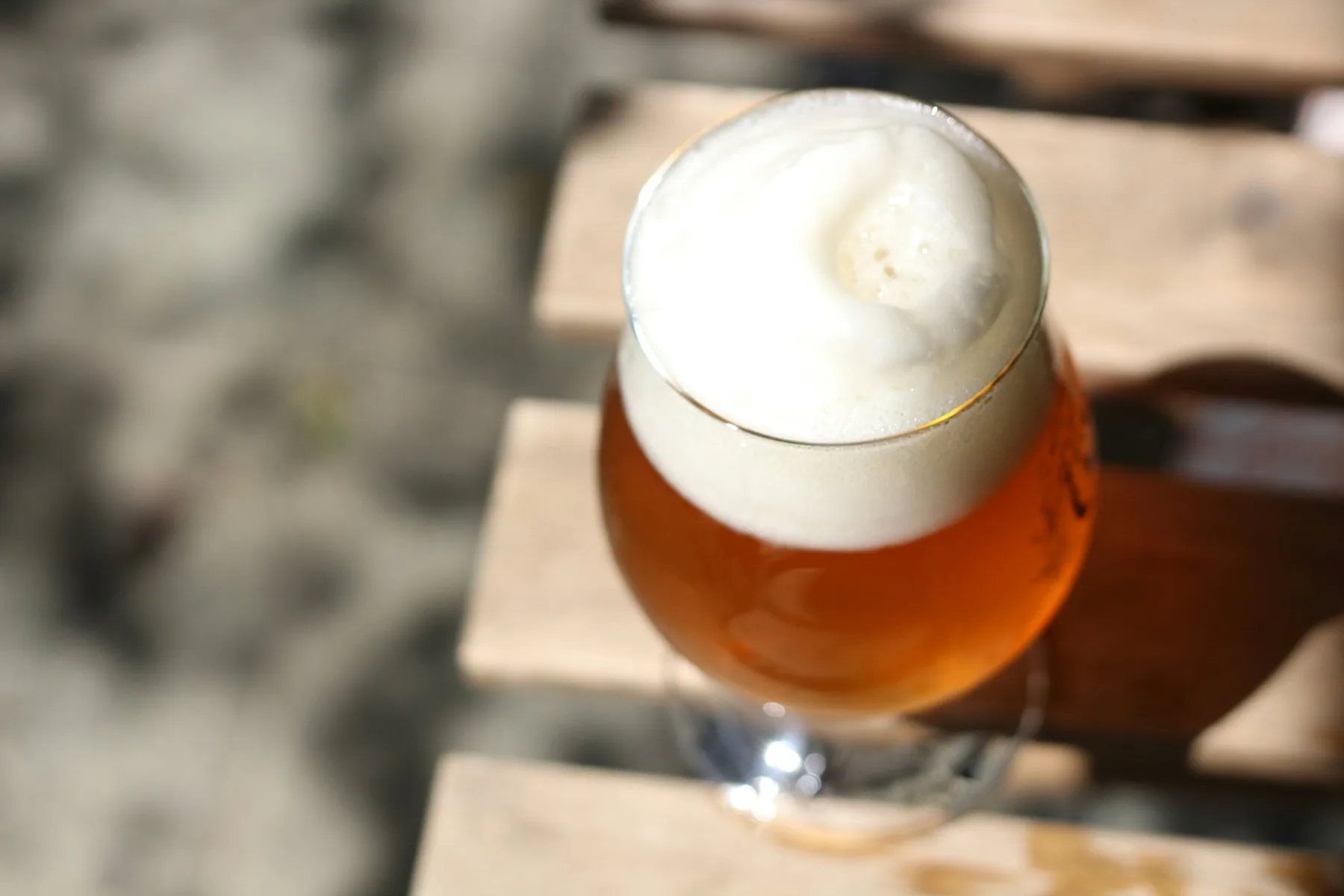 Et tulipanformet glass med ravgult ol og tykt, hvitt skum står på et bord av lyse trelemmer i skarpt sollys.