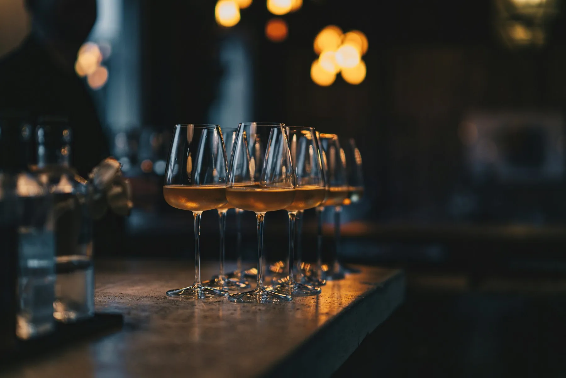 En rekke like vinglass med en liten slant lys vin står tett sammen på en bardisk, med varme lyspunkter ute av fokus i mørket bak.