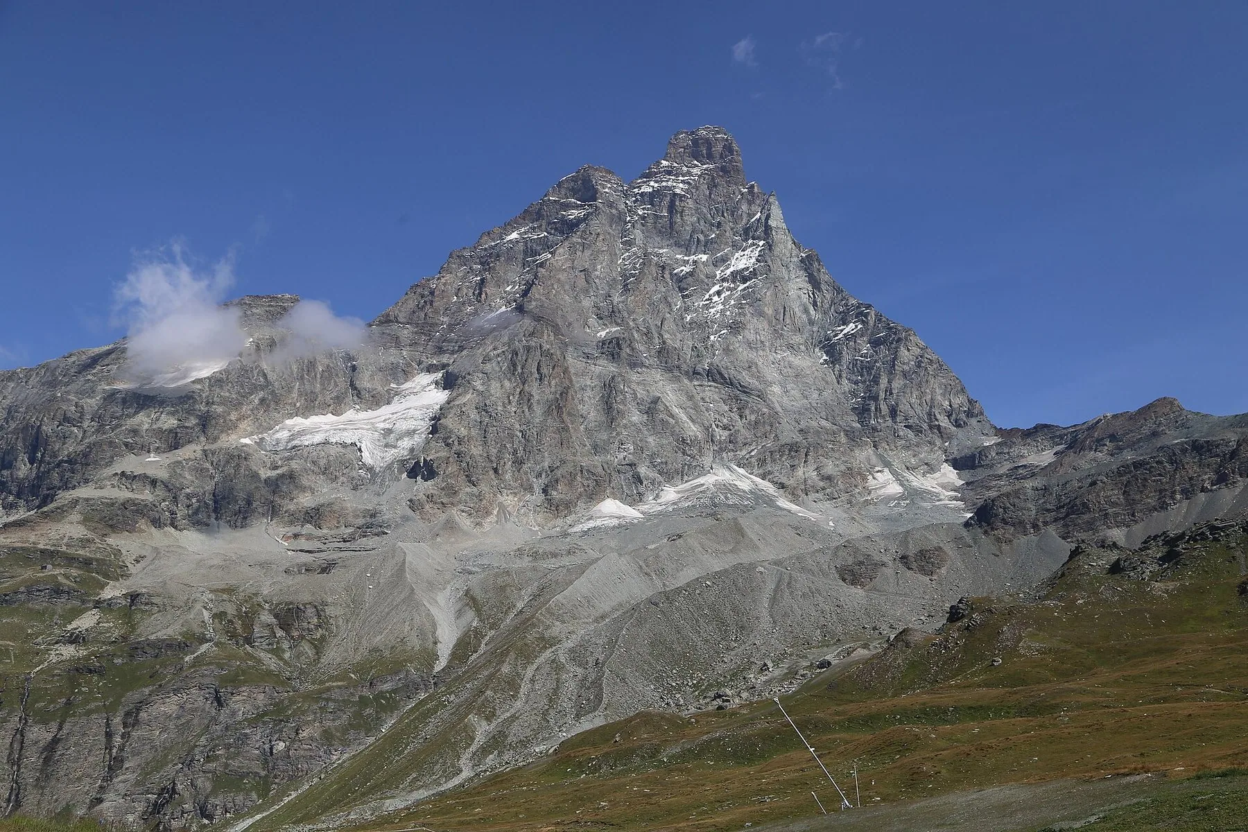 Et bratt, pyramideformet fjell med snøkledde flanker og en bre nedenfor, sett fra sørsiden mot en skyfri blå himmel.